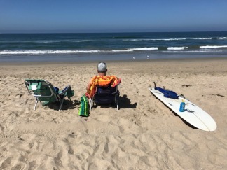 A man sitting at the beach with a beach chair and surfboard.